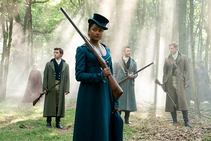 lady standing in the woods in period clothing, holding a gun, three men are standing behind her